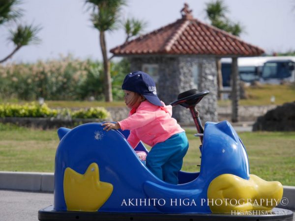 子供が青い乗用玩具に乗り、公園で遊ぶ様子の写真。
